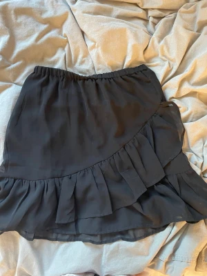 Svart volangkjol Pernilla Wahlgren - Supersnygg svart kortkjol från Pernilla Wahlgren med volangdetaljer och elastisk midja. Kjolen har en lätt och flowy känsla, perfekt att styla med en cool topp eller stickad tröja. Materialet känns mjukt och följsamt.