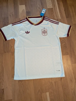 Vit Spanien fotbollströja Adidas - .säljer en Vit Spanien fotbollströja från Adidas den har lite stor krage men annars fin.  med vinröda och guldiga detaljer. Tröjan har korta ärmar, broderad landslagssköld och Adidas-logga på bröstet samt klassiska ränder på axlarna. Tillverkad i lätt och ventilerande material, perfekt för träning eller match.