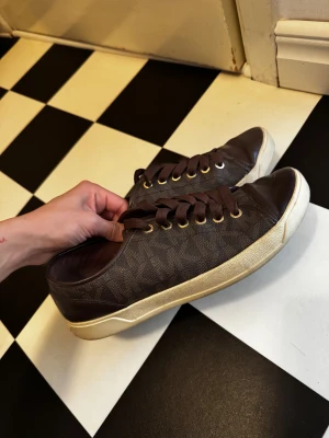 Bruna Michael Kors sneakers - Snygga bruna sneakers från Michael Kors med klassiskt MK-mönster på sidan, bruna skosnören och guldfärgade öljetter. Ovandel i textil och läderdetaljer, vit sula som ger en fräsch kontrast. Perfekta för en trendig och avslappnad look.