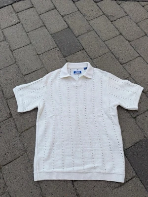 Vit stickad pikétröja från Jaxon - Vit stickad pikétröja från Jaxon med korta ärmar och klassisk krage. Tröjan har ett diskret hålmönster över hela plagget och ribbstickade muddar vid ärmslut och nederkant. Perfekt för en stilren och avslappnad look.