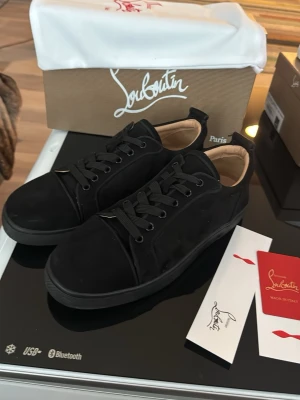 Svarta Louboutin sneakers med röd sula - svarta sneakers från Christian Louboutin med klassisk röd sula och svarta skosnören. Ovandelen är i mocka och insidan har en beige läderdetalj. Perfekta för dig som vill ha en lyxig och tidlös look.