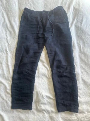 Mörkblå linnebyxor med snöre  - Snygga mörkblå linnebyxor med avslappnad passform och snören i midjan. Byxorna har raka ben och sidofickor, perfekta för en chill och stilren look. Tillverkade i svalt och skönt linne som passar bra till varmare dagar. Storlek L