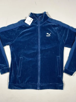 Blå velourtröja  Puma (finns även i storlek S) - Mysig mörkblå velourtröja från Puma med dragkedja hela vägen, krage och två fickor framtill. Tröjan har en sportig retro look men avslappnad. Logga på bröstet ton-i-ton paneler längs ärmarna. Perfekt för en sportig men trendig stil.