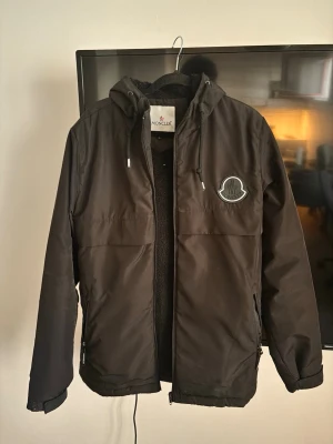 Svart vindjacka från Moncler - Svart vindjacka från Moncler med huva och dragkedja. Jackan har en stor logotyp på bröstet och är fodrad med mjukt material på insidan. Praktiska fickor och justerbar huva gör den perfekt för blåsiga dagar. Dragkedjan har defekt, men annars är den i bra skick.