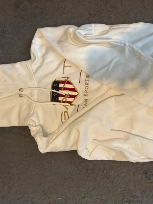 Vit hoodie från Gant med tryck - Snygg vit hoodie från Gant med klassiskt tryck och sköld på bröstet. Hoodien har dragsko i huvan och en stor magficka. Perfekt för dig som gillar en clean och sportig stil. Materialet känns mjukt och bekvämt.