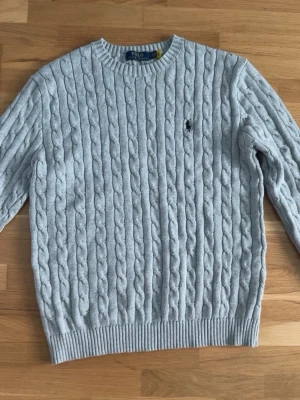 Ljusgrå kabelstickad tröja Polo Ralph Lauren - Snygg ljusgrå kabelstickad tröja från Polo Ralph Lauren med rund halsringning och broderad logga på bröstet. Tröjan har ribbade muddar vid ärmslut och nederkant. Perfekt för dig som gillar klassisk och stilren design.