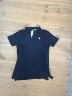 Svart pikétröja från Abercrombie - Klassisk svart pikétröja från Abercrombie & Fitch med liten logga på bröstet. Tröjan har krage, knappar framtill och korta ärmar. Perfekt till jeans eller shorts för en avslappnad look.