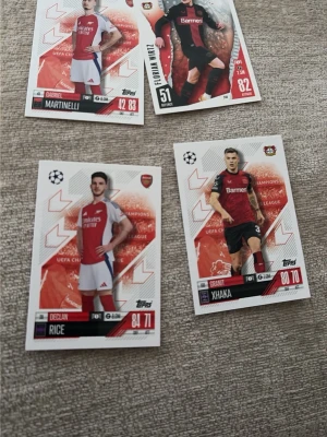 Fyra Topps fotbollssamlarbilder - Säljer fyra fotbollssamlarbilder från Topps med spelare från Arsenal och Bayer Leverkusen. Korten har vit bakgrund med röda och svarta detaljer, spelarnas namn och lagloggor. Perfekt för dig som gillar att samla på fotbollskort och vill utöka din samling.