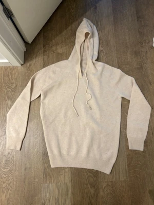 Beige ull/merinoull/Kashmir  - Säljer en stilren beige hoodie med huva och dragsnören. Hoodien har långa ärmar och ribbade muddar vid ärmslut och nederkant. Perfekt för en avslappnad look och supermjuk mot huden. Vet ej riktigt vad det är för tyg. 