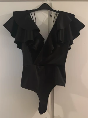 Svart volangbody  - Snygg svart body från Gina med djup V-ringning och stora volangdetaljer över axlarna. Tillverkad i ett slätt, glansigt material som ger en lyxig känsla. Perfekt för dig som vill ha något extra till outfiten. Modellen har smala axelband och markerad midja.
