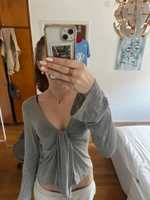 Grå omlottopp från Gina Tricot - Supersnygg grå omlottopp från Gina Tricot med långa ärmar och knyt framtill. Toppen har en djup v-ringning och är tillverkad i mjuk bomullsblandning. Perfekt för en trendig och avslappnad look. Passar mig som har S/M