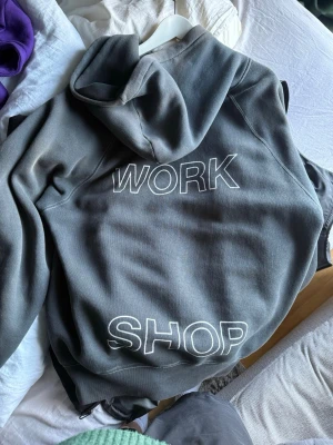 Grå hoodie från Workshop - Snygg grå hoodie från Our Legacy med stor vit text 'WORK SHOP' på ryggen och en vit grafisk logga på bröstet. 