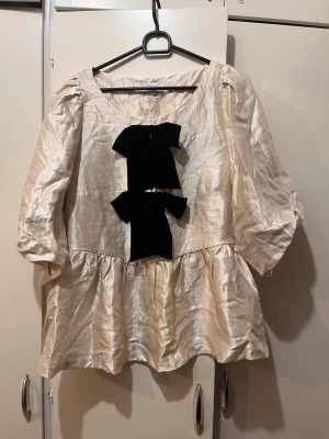 Peplum Blus guld beige Joelle strl 40  - Peplum topp från Joelle i strl 40. Färgen är skimrande guld bege cremefärgad. Avtagbara rosetter framtill, puffärmar och volang nertill. Helt ny, aldrig använd. Köpt från Ellos. Nypris: 699kr 