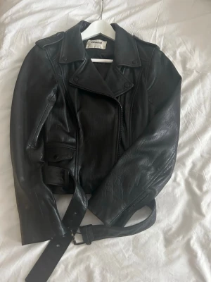Svart skinnjacka från Stand - Snygg svart skinnjacka från Stand med klassisk bikerstil, breda slag, sned dragkedja och bälte i midjan. Jackan har fickor med lock och tryckknappar samt axelklaffar, allt i svarta detaljer. Skinnet är väldigt mjukt och följsamt. Sparsamt använd och nu för liten tyvärr. Pris på Vestiare är ca 3000kr. 
