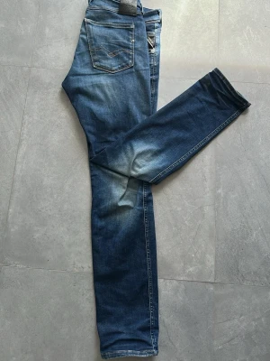 Blå slim fit jeans från Replay - Snygga blå slim fit jeans från Replay med klassisk femficksdesign och diskreta slitningar på låren och knäna. Jeansen har normal midja och raka ben, tillverkade i stretchigt denim för extra komfort. Perfekta för en avslappnad och trendig look.