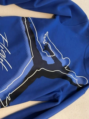 Blå Jordan Flight hoodie - Snygg blå hoodie från Jordan med stort Flight-tryck och ikonisk siluett på framsidan. Hoodien har långa ärmar och en sportig vibe, perfekt för dig som gillar streetwear och basketstil. Materialet känns mjukt och bekvämt mot huden.