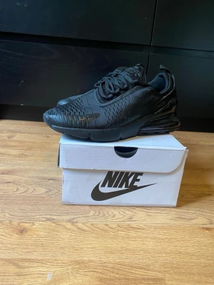 Nike Air Max 270 Triple Black - Snygga Nike Air Max 270 i helsvart färg. Skorna har en chunky sula med synlig Air-enhet i hälen, mesh- och syntetmaterial på ovandelen samt klassisk Nike-logga på sidan. Perfekta för dig som gillar streetwear och sportig stil.