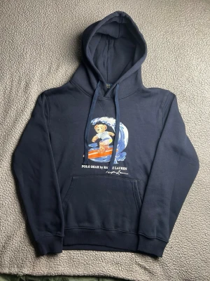 Marinblå hoodie Polo Ralph Lauren - Snygg marinblå hoodie från Polo Ralph Lauren med den ikoniska Polo Bear som surfar på bröstet. Mycket bra skick. Skickar snabbt efter att du har beställt. Frågor? Skicka ett meddelande.