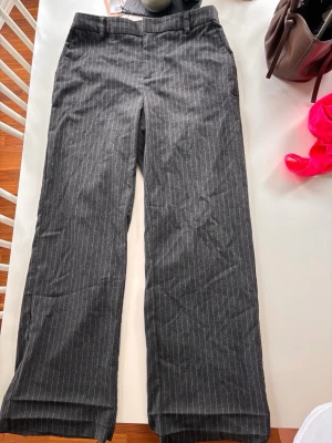Grå  straight/bootcut kostymbyxor med ränder Gina Tricot - Snygga grå lågmidjade kostymbyxor från Gina Tricot med vita tunna kritstreck. Byxorna har raka/löst bootcut ben, hällor för skärp och klassisk dragkedja fram. Avklippta längst ner för att passa någon som är runt 160cm som man enkelt kan sy snyggare/använda klisterfåll.