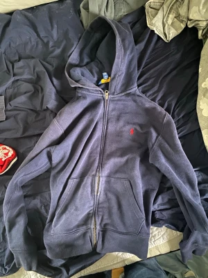 Marinblå hoodie från Polo Ralph Lauren - Snygg marinblå hoodie med dragkedja från Polo Ralph Lauren. Klassisk design med röd broderad logga på bröstet, huva och kängurufickor. Mjuk bomullskänsla och perfekt för en chill och stilren look.
