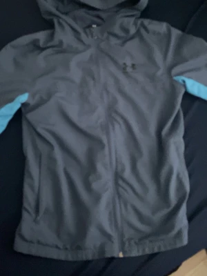 Blå vindjacka från Under Armour - Snygg grå vindjacka från Under Armour med huva och dragkedja framtill. Jackan har ljusblå detaljer längs ärmarna och två fickor på sidorna. Perfekt för blåsiga dagar och sportiga outfits.