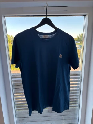 Mörkblå Moncler t-shirt - Snygg mörkblå t-shirt från Moncler med klassisk rund halsringning och liten Moncler-logga på bröstet. Tillverkad i mjuk bomull som känns skön mot huden. Perfekt för en stilren och enkel look. Står att det är storlek L men passar M lika bra.