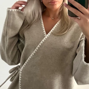 Beige omlottkofta - Beige omlottkofta i mjukt material med knytband i midjan och dekorativ vit kant längs öppningen. Koftan har vida ärmar och en avslappnad passform som ger en chill och trendig look.