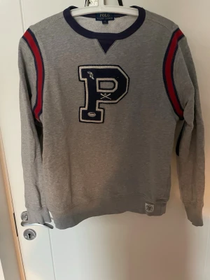 Grå sweatshirt Polo Ralph Lauren - Snygg grå sweatshirt från Polo Ralph Lauren med stort P-tryck på bröstet och blå samt röda ränder på axlarna. Tröjan har rund halsringning med blå kant och är tillverkad i mjukt bomullsmaterial. Perfekt för en sportig och avslappnad stil.