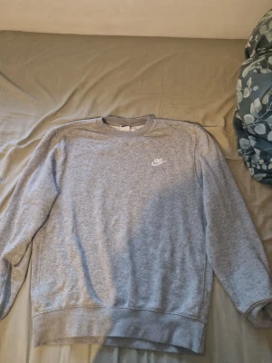 Grå sweatshirt från Nike - Klassisk grå sweatshirt från Nike med rund halsringning och diskret Nike-logga i vitt på bröstet. Tröjan har ribbade muddar vid ärmslut och nederkant, perfekt för en avslappnad och sportig stil.
