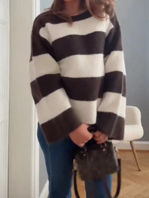 Randig stickad tröja - Säljer en stickad tröja med breda bruna och vita ränder. Tröjan har en avslappnad passform, rund halsringning och långa ärmar. Perfekt för en chill och trendig look.