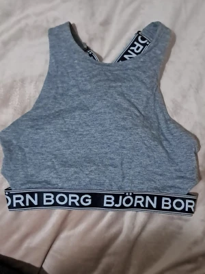 Grå sport-bh från Björn Borg - Sportig grå bh från Björn Borg med bred resår och logotypdetaljer i svart och vitt. Modellen har hög halsringning och korsade axelband i ryggen för extra stöd. Tillverkad i mjukt och stretchigt material som ger en bekväm passform. Liten i storlek