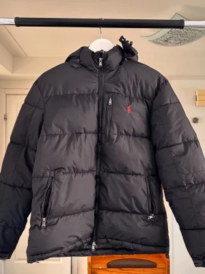 Svart pufferjacka från Ralph Lauren - Snygg svart pufferjacka från Ralph Lauren med huva och dragkedja framtill. Jackan har två stora fickor med dragkedja och den klassiska röda Polo-loggan broderad på bröstet. Perfekt för kalla dagar och riktigt skön att bära. Materialet är slitstarkt och håller värmen bra.