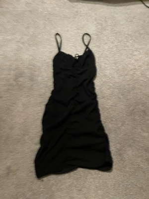 Svart kort klänning med smala band - Säljer en svart kort klänning med smala axelband och snörning framtill. Klänningen har en enkel, figurnära passform och är tillverkad i ett lätt, strukturerat tyg. Perfekt för dig som gillar minimalistisk stil. Köpt på New Yorker.