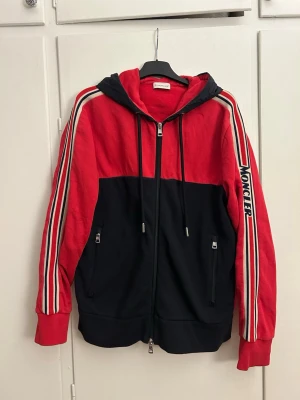 Röd och svart hoodie från Moncler - Snygg hoodie från Moncler i rött och svart med dragkedja framtill, två fickor med dragkedja och huva. Ränder i vitt, rött och blått längs ärmarna samt Moncler-logga på vänster ärm. Vattenavvisande luva. Fint skick. L men sitter mer som M