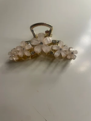 Hårklämma med blommor i guld och rosa - Söt hårklämma i guldfärgad metall dekorerad med ljusrosa blommor i plast och små glittriga stenar i mitten. Klämman har en elegant form och fina detaljer på både fram- och baksidan. Perfekt accessoar för att piffa upp frisyren.