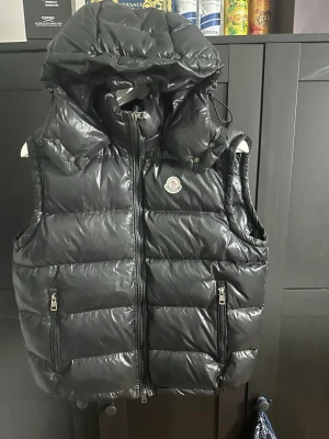 Svart dunväst från Moncler - Snygg svart dunväst från Moncler med glansig finish och stor huva. Västen har två framfickor med dragkedja och Moncler-logga på bröstet. Perfekt för lager-på-lager och håller dig varm med sin fluffiga dunfyllning. Size 2