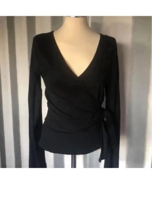 Svart omlottopp med knytband - Stilren svart omlottopp med långa ärmar och v-ringning. Toppen har knytband i sidan som ger en snygg passform och markerar midjan. Perfekt att matcha med jeans eller kjol för en trendig look.