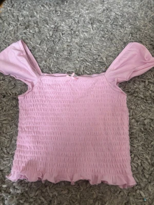 Ljusrosa smockad topp med volang - Söt ljusrosa topp med smockad struktur och volangkant. Toppen har korta ärmar som kan bäras offshoulder och en liten rosett framtill. Perfekt för dig som gillar romantiska och trendiga plagg.