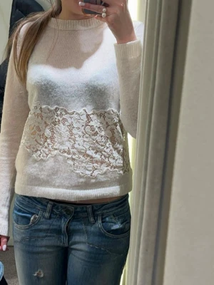 Vit stickad tröja med spetsdetalj - Trendig vit stickad tröja med bred spetsdetalj över magen. Tröjan har rund halsringning och långa ärmar. Spetsen har blommigt mönster över magen. Tröjan är i storlek S, men är något liten i storleken.