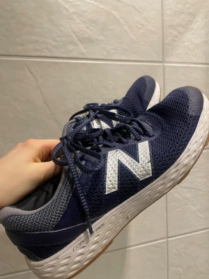 Marinblå New Balance sneakers - Snygga marinblå sneakers från New Balance med vit sula och stort vitt N på sidan. Ovandelen är i mesh för bra ventilation och skorna har klassiska snören. Perfekta för dig som gillar sportig stil och vill ha bekväma skor till vardags.