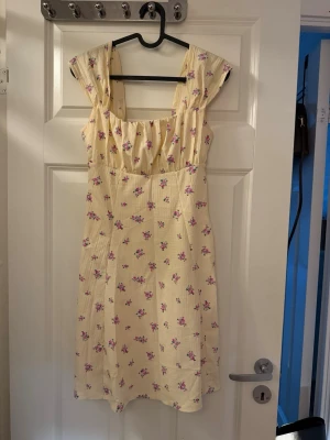 Blommig ljusgul sommarklänning - Söt ljusgul klänning med små rosa och lila blommor. Klänningen har breda axelband, rynkad byst och är tillverkad i ett lätt, strukturerat bomullstyg. Perfekt för varma dagar och har en feminin, avslappnad passform.