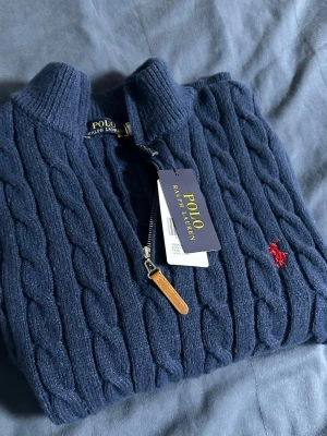 Mörkblå kabelstickad tröja Polo Ralph Lauren - Kabelstickad mörkblå tröja från Polo Ralph Lauren med halv dragkedja och klassisk röd broderad logga på bröstet. Tröjan har hög krage och detaljer i läder på dragkedjan. Perfekt för dig som gillar stilren och tidlös design.