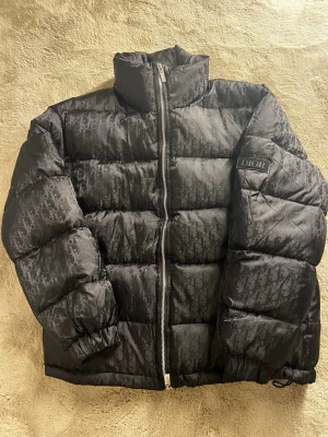 Svart pufferjacka från Dior - Säljer en svart pufferjacka från Dior med ikoniskt monogrammönster över hela jackan. Jackan har hög krage, dragkedja framtill och logotyp på ärmen. Perfekt för kalla dagar och riktigt snygg att bära till jeans eller joggers.