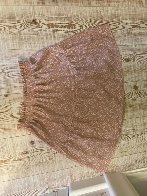 Kort blommig kjol med volang - Söt kort kjol i ljusbrun ton med små vita blommor och volangkant. Kjolen har bred resår i midjan för skön passform och är gjord i mjukt bomullstyg. Storlek S men justerbar i midjan. Hör av er om ni vill ha bilder, priset kan alltid diskuteras!