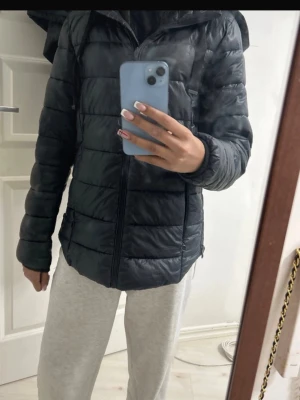 Svart pufferjacka med huva - Säljer en svart pufferjacka med huva och dragkedja framtill. Jackan har två sidofickor med dragkedja och quiltad design. Perfekt för kalla dagar och ger en stilren look. Materialet är glansigt och lätt vadderat för extra värme.