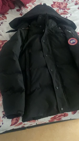 Svart dunjacka från Canada Goose - Säljer en svart dunjacka från Canada Goose med huva och klassisk logga på ärmen. Jackan har lång ärm, knappar och dragkedja framtill. Perfekt för kalla dagar med sin tjocka och puffiga design. Stilsäker och funktionell för vintern.