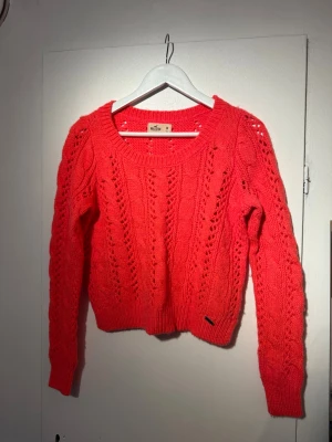 Stickad tröja från Hollister - Snygg korallröd stickad tröja från Nelly med hålmönster och bred ribbad kant vid hals, ärmslut och nederkant. Tröjan har en kortare passform och rund halsringning. Perfekt för att ge din outfit lite färg.