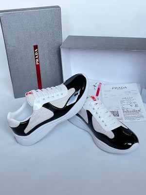 Svarta och vita sneakers från Prada - Snygga sneakers från Prada i svart och vitt med glansiga svarta partier och meshdetaljer. Klassisk låg modell med vit sula och snörning. Diskret röd Prada-logga på plösen ger en exklusiv touch. Perfekta för dig som vill ha en stilren och modern look.