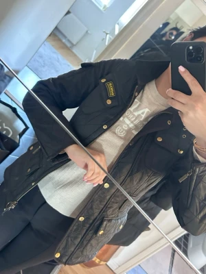 Svart quiltad jacka från Barbour - Snygg svart quiltad jacka från Barbour med guldiga knappar och dragkedja. Klassisk modell med flera fickor och axelklaffar. Jackan har en lätt figursydd passform och är perfekt för dig som vill ha en stilren och tidlös look.