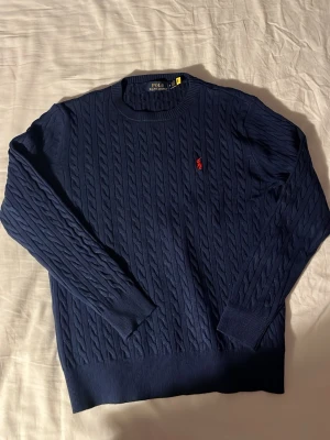 Mörkblå  stickadtröja Polo Ralph Lauren - Kabelstickad mörkblå tröja från Polo Ralph Lauren med klassisk röd logga broderad på bröstet. Tröjan har rund halsringning, långa ärmar och ribbade muddar. Perfekt för dig som gillar tidlös och stilren design.